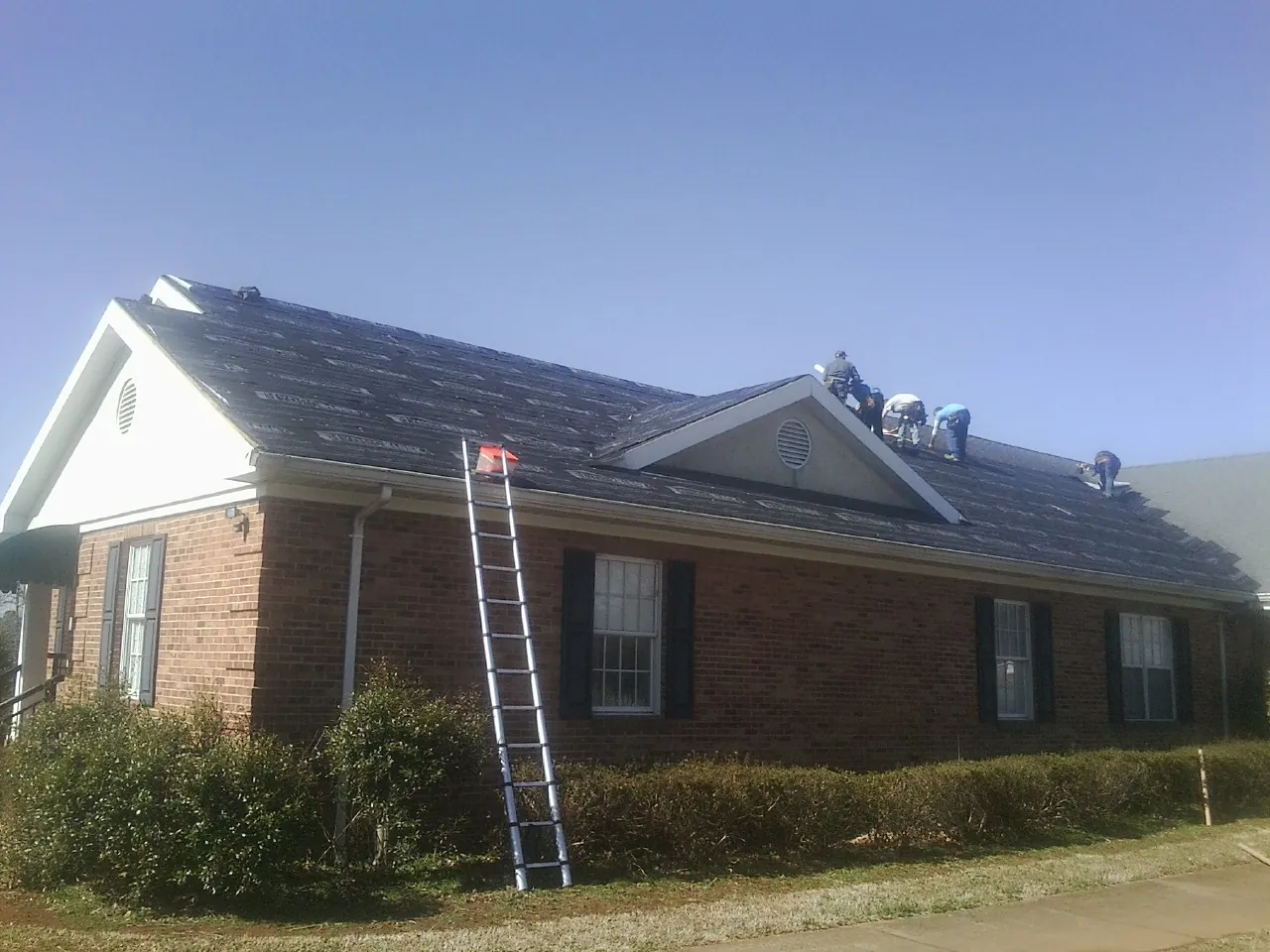 Crew tearing shingles off a brick ranch roof with a ladder set — Metal Roof Repair work by our crew in Hector